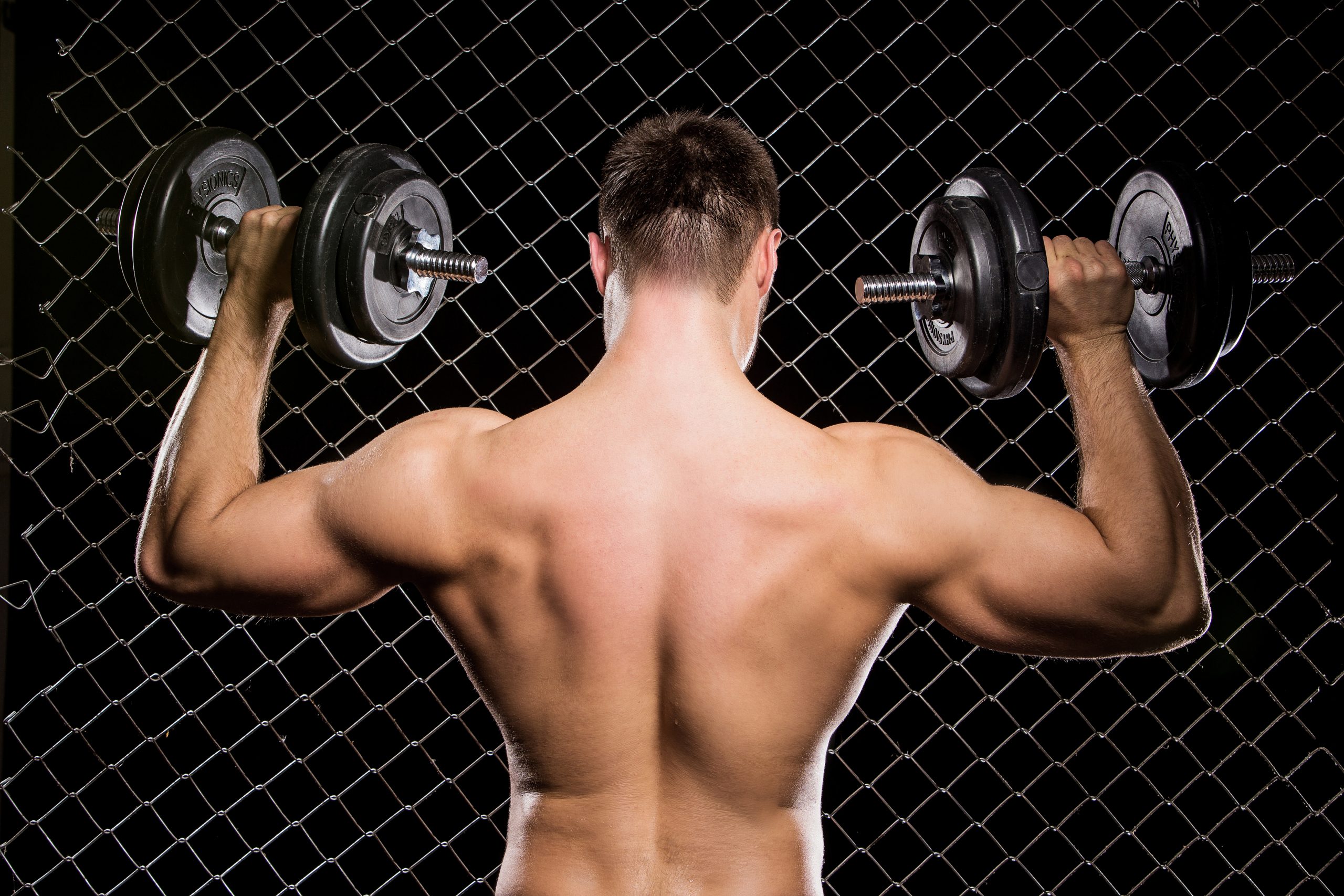 Powerful guy with a dumbbells showing muscles on fence backgroun